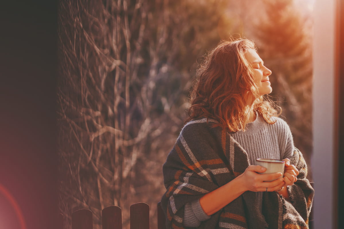 Frau mit Teetasse in der Hand, die in der Herbstsonne steht, mit geschlossenen Augen.